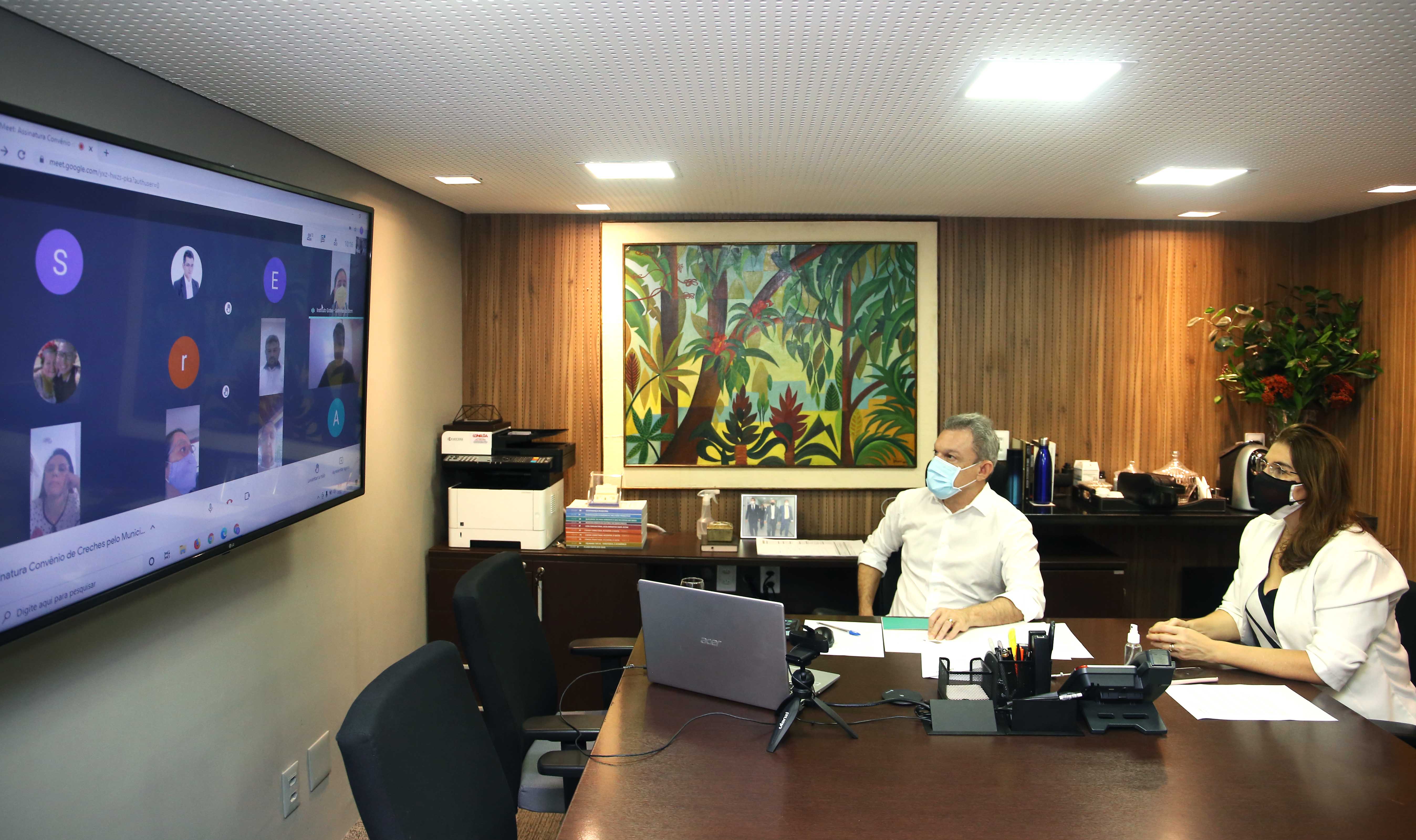 prefeito sarto e secretária dalila sentado em uma mesa de reunião diante de um telão, que mostra as janelas da reunião virtual